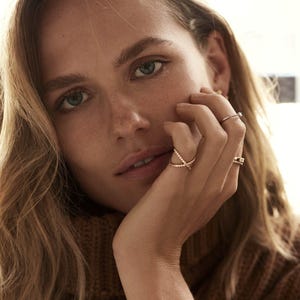May include: A close-up shot of a person's hand adorned with several rings. The rings feature delicate designs, including a crisscross style and a band with a small stone. The person is wearing a brown sweater.