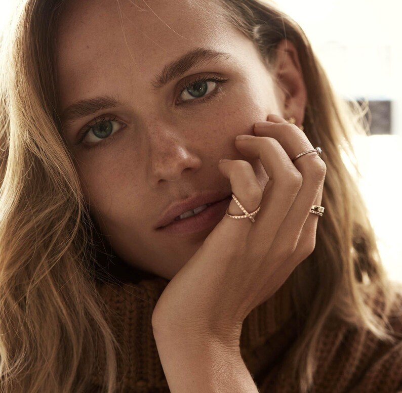 May include: A close-up shot of a person's hand adorned with several rings. The rings feature delicate designs, including a crisscross style and a band with a small stone. The person is wearing a brown sweater.
