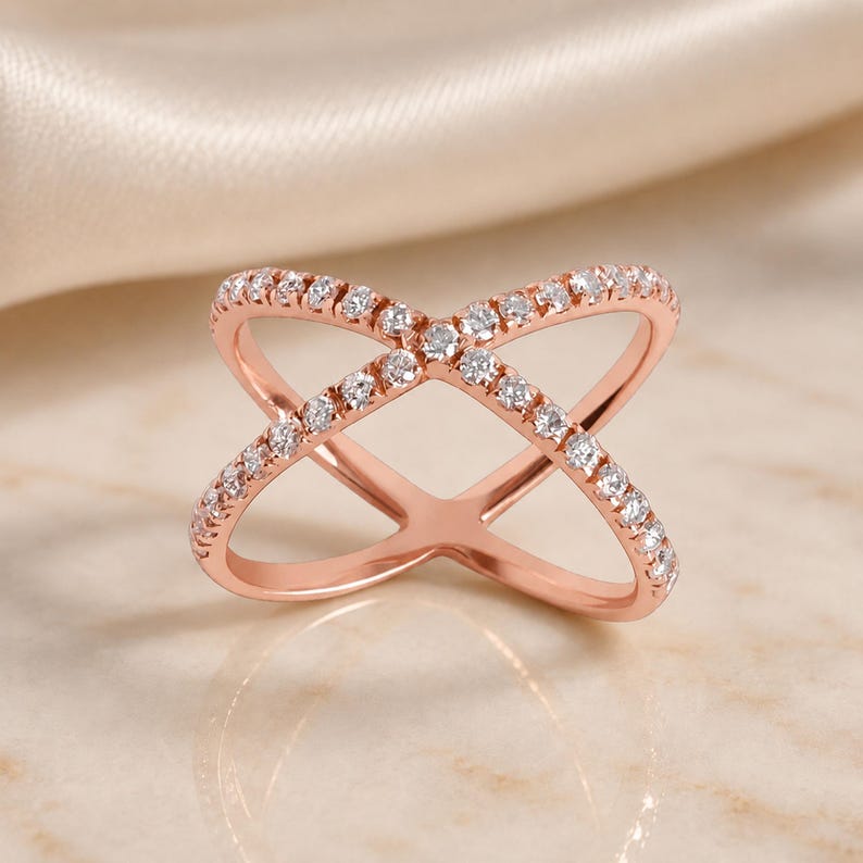 May include: Rose gold criss-cross ring with a double band design. The ring is encrusted with small, clear gemstones along the bands. The ring is displayed on a light-colored surface.