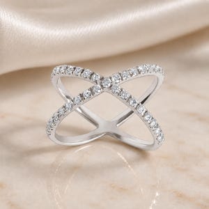 May include: A silver-toned ring with a crisscross design, encrusted with small, sparkling clear stones. The ring's band forms an X shape, with the stones set along the top of the band. The ring is displayed on a light-colored surface.