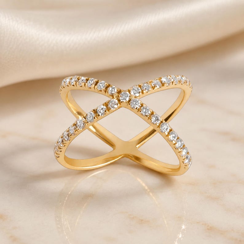 May include: A gold-colored ring with a crisscross design, encrusted with small, clear gemstones. The ring's band is a continuous loop, and the gemstones are evenly spaced along the crisscrossing bands. The ring is displayed on a light-colored surface.