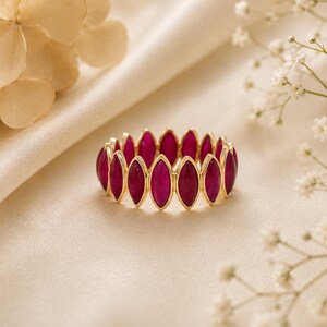 May include: A gold ring featuring a series of oval-shaped, deep red gemstones. The gemstones are set closely together, creating a continuous band of color. The ring is set against a soft, cream-colored background, with delicate white flowers in the background.