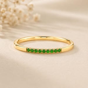Puede incluir: Un anillo de banda dorado con una fila de pequeñas piedras preciosas verdes vibrantes. El anillo tiene un acabado liso y pulido y un diseño delicado. Las piedras preciosas están espaciadas uniformemente en la parte superior del anillo.