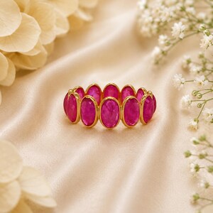 Könnte beinhalten: Ein goldener Ring mit ovalen, leuchtend rosa Edelsteinen. Die Edelsteine sind eng aneinander gesetzt und bilden ein durchgehendes Farbband. Der Ring wird auf einer cremefarbenen Satin-Oberfläche mit floralen Akzenten im Hintergrund präsentiert.