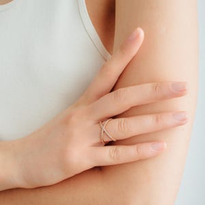 May include: A rose gold ring with a crisscross design, encrusted with small, sparkling stones. The ring is shown on a hand, highlighting its delicate and elegant features. This piece of jewelry is suitable for everyday wear or special occasions.