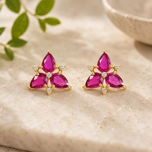 Puede incluir: Un par de pendientes de oro. Cada pendiente presenta un diseño floral con tres gemas de color magenta en forma de lágrima y pequeñas gemas transparentes. Los pendientes están colocados sobre un fondo neutro.