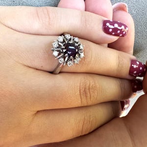 Puede incluir: Un anillo de plata con una piedra preciosa de color púrpura oscuro rodeada de pequeñas piedras preciosas claras. El anillo se muestra sobre un fondo color piel. El diseño del anillo es un grupo de piedras, creando un efecto floral o de estrella.