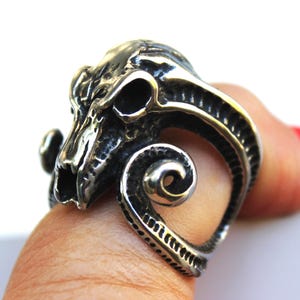 Könnte beinhalten: Ein silberfarbener Ring mit detailliertem Widder-Schädel-Design. Der Ring hat eine strukturierte Oberfläche und ein markantes, gebogenes Horn. Die Augenhöhlen und Gesichtszüge des Schädels sind klar definiert, was eine gotische oder Biker-Ästhetik erzeugt.