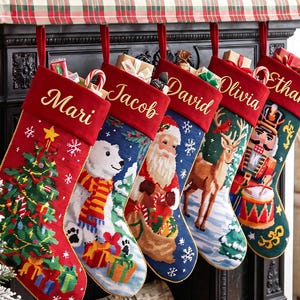 May include: Five needlepoint Christmas stockings hanging in a row, each featuring a red cuff with a personalized name in gold script. The stockings display festive scenes including a decorated tree, a polar bear, Santa Claus, a reindeer, and a nutcracker.