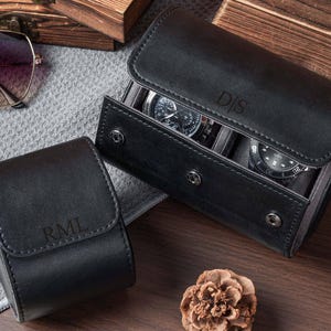 May include: Two black leather watch cases with snap closures. One smaller case features the embossed initials RML, while the larger case holds two wristwatches and is embossed with the initials D|S on the front flap.