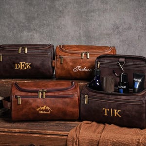 May include: Several brown leather wash bags with gold zips and embroidered initials or names. One bag is open, displaying grooming products. The bags are on a wooden surface with grooming tools.