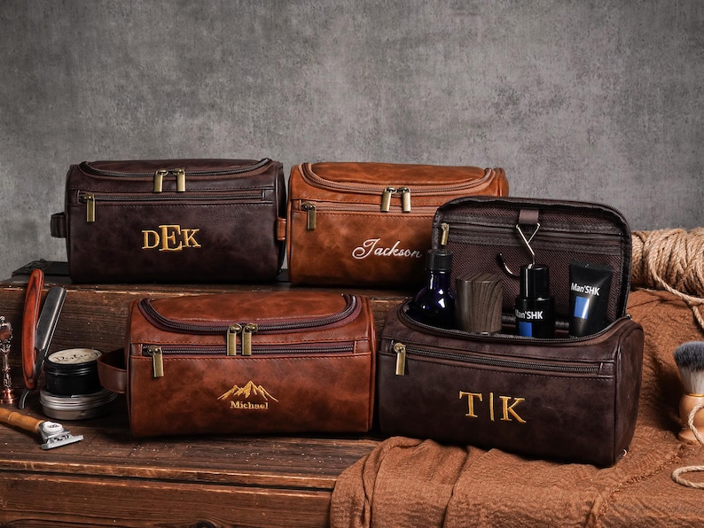 May include: Several brown leather wash bags with gold zips and embroidered initials or names. One bag is open, displaying grooming products. The bags are on a wooden surface with grooming tools.