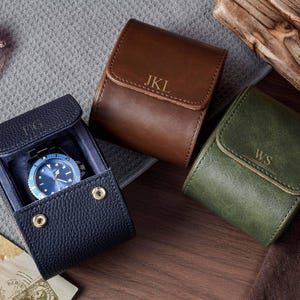 May include: Three leather watch cases in navy blue, brown, and olive green. The navy case is open, revealing a blue-faced wristwatch inside. Each case features a flap with a gold-tone snap closure and personalized initials embossed on the front.