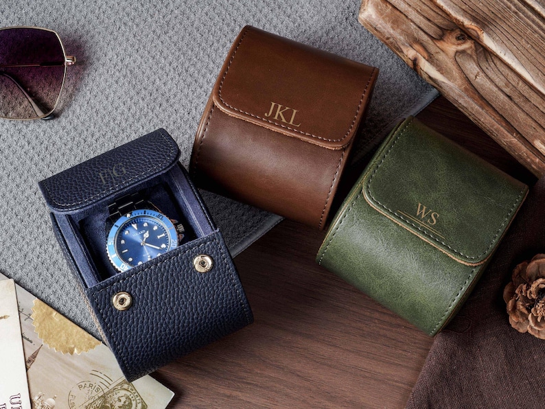May include: Three leather watch cases in navy blue, brown, and olive green. The navy case is open, revealing a blue-faced wristwatch inside. Each case features a flap with a gold-tone snap closure and personalized initials embossed on the front.