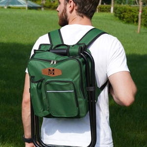 May include: A forest green backpack with an integrated black metal folding stool frame. The bag features multiple zippered compartments, a side pocket, and a tan leather-like patch on the front with the black text "MICHAEL" and a letter M.
