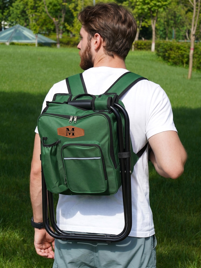 May include: A forest green backpack with an integrated black metal folding stool frame. The bag features multiple zippered compartments, a side pocket, and a tan leather-like patch on the front with the black text "MICHAEL" and a letter M.