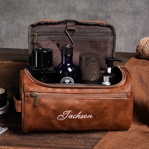 May include: A brown leather wash bag, embroidered with the name "Jackson", open to reveal grooming products. These include a dark blue bottle, a wooden cup, and a shaving brush. A straight razor and shaving cream are also visible.