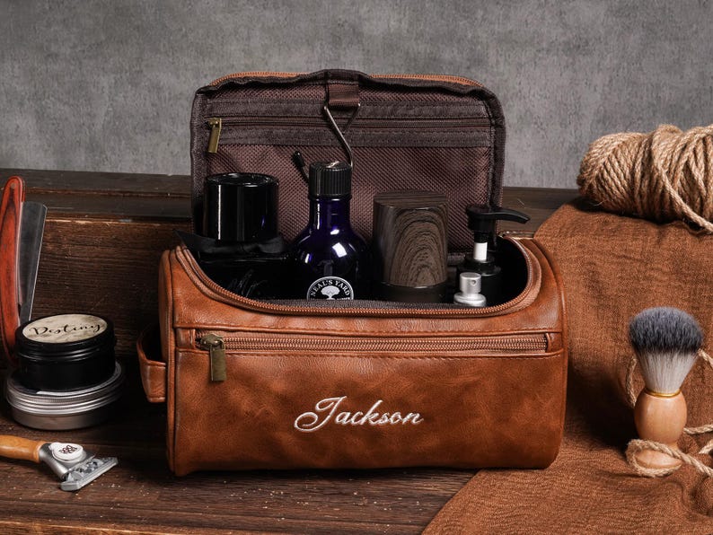 May include: A brown leather wash bag, embroidered with the name "Jackson", open to reveal grooming products. These include a dark blue bottle, a wooden cup, and a shaving brush. A straight razor and shaving cream are also visible.