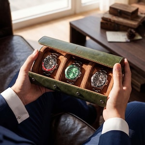 May include: A forest green leather watch roll case with a tan interior, designed to hold three wristwatches. The case features a snap-button closure and contains three silver-toned watches with black, green, and red-accented circular faces.