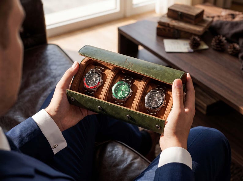 May include: A forest green leather watch roll case with a tan interior, designed to hold three wristwatches. The case features a snap-button closure and contains three silver-toned watches with black, green, and red-accented circular faces.