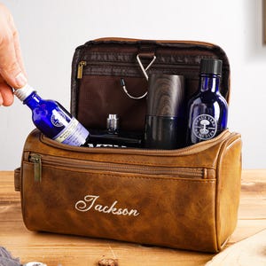 May include: A brown leather wash bag with the name "Jackson" embroidered on the front. The bag is open, showing blue bottles and a wooden container. A hand is placing a blue bottle into the bag.
