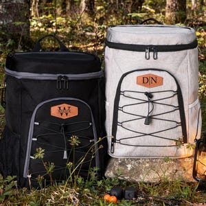 May include: Two insulated cooler backpacks, one black and one light gray, with black accents and a front pocket with a decorative cord. Each has a leather patch with initials. A lit lantern sits nearby, suggesting outdoor use.
