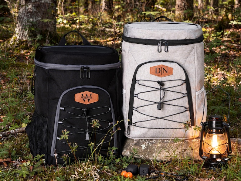 May include: Two insulated cooler backpacks, one black and one light gray, with black accents and a front pocket with a decorative cord. Each has a leather patch with initials. A lit lantern sits nearby, suggesting outdoor use.