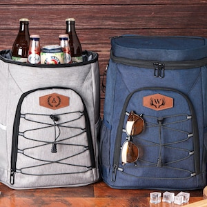 May include: Two insulated backpack coolers, one gray and one navy blue, with black accents. The gray cooler has a leather patch with the letter "A", and the blue cooler has a leather patch with a deer antler design. Both have a bungee cord system.