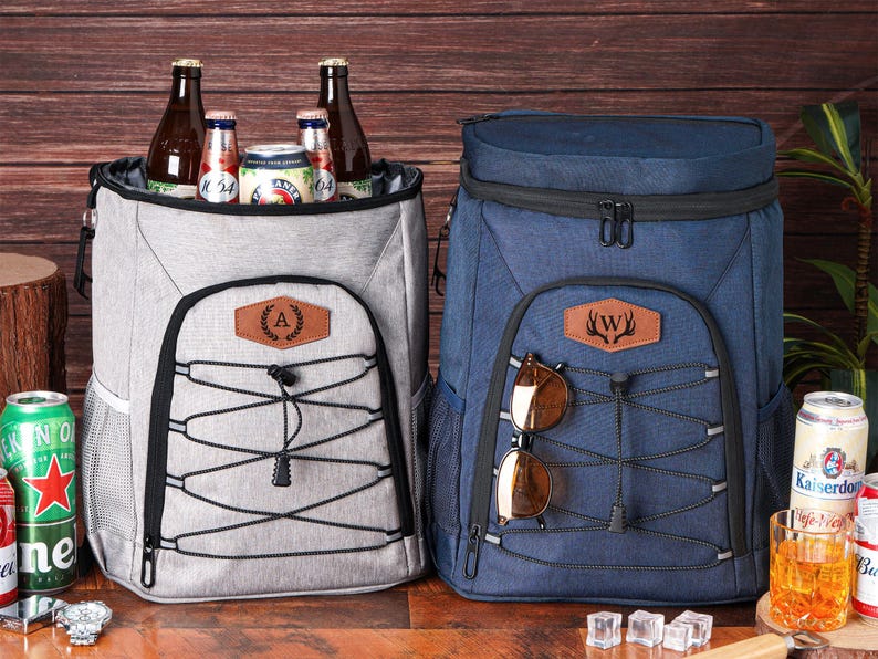 May include: Two insulated backpack coolers, one gray and one navy blue, with black accents. The gray cooler has a leather patch with the letter "A", and the blue cooler has a leather patch with a deer antler design. Both have a bungee cord system.