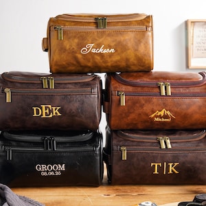 May include: A collection of personalised leather wash bags in shades of brown and black. Each bag has a zip closure and is embossed with a name or initials in gold lettering. The bags are displayed on a wooden surface.