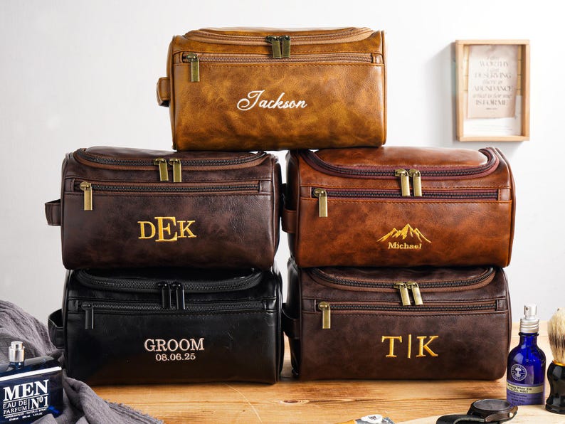 May include: A collection of personalised leather wash bags in shades of brown and black. Each bag has a zip closure and is embossed with a name or initials in gold lettering. The bags are displayed on a wooden surface.