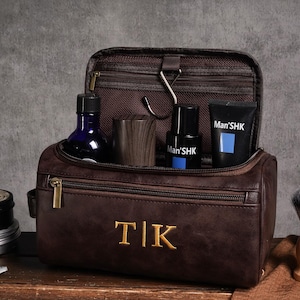 May include: A brown leather wash bag with gold zip and embroidered initials. Inside are grooming products, including a blue glass bottle, wooden cup, and black tubes with blue accents. A shaving brush and razor are also visible.