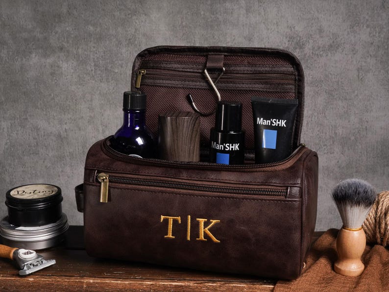 May include: A brown leather wash bag with gold zip and embroidered initials. Inside are grooming products, including a blue glass bottle, wooden cup, and black tubes with blue accents. A shaving brush and razor are also visible.