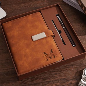 Puede incluir: Un juego de diario y bolígrafo de cuero marrón en una caja de regalo. El diario tiene un cierre plateado y el nombre "Martin" grabado en la portada. Se incluye un bolígrafo plateado y un lápiz óptico. Se ve un reloj.