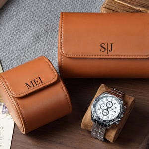 May include: Two tan leather watch cases with rounded flaps and magnetic closures. One case is engraved with the letters SJ, and the other is engraved with the name MEI. A silver-toned chronograph wristwatch with a white face rests on a tan cushion.