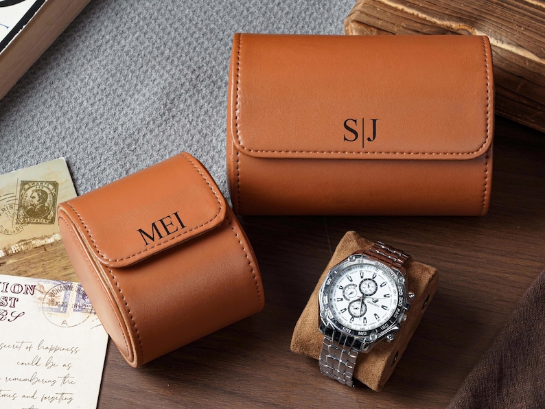 May include: Two tan leather watch cases with rounded flaps and magnetic closures. One case is engraved with the letters SJ, and the other is engraved with the name MEI. A silver-toned chronograph wristwatch with a white face rests on a tan cushion.