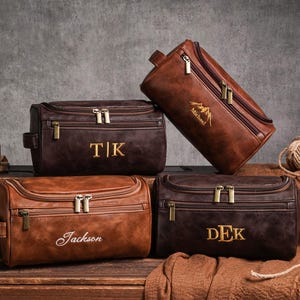 May include: Four brown leather wash bags with gold zips and personalised text. One bag is tilted, displaying a mountain graphic and the name "Michael". Other bags feature initials or names like "TK", "Jackson", and "DEK".