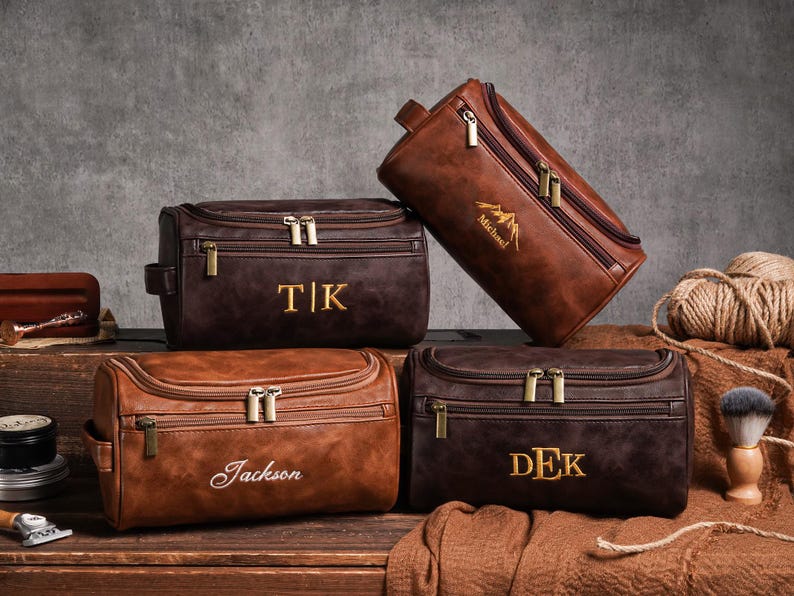 May include: Four brown leather wash bags with gold zips and personalised text. One bag is tilted, displaying a mountain graphic and the name "Michael". Other bags feature initials or names like "TK", "Jackson", and "DEK".