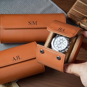 May include: Three tan leather watch rolls with snap closures and personalized monograms. One roll is open, revealing a silver-toned chronograph wristwatch with a white face inside. The monograms S|M, AJL, and BA are embossed on the exterior flaps.