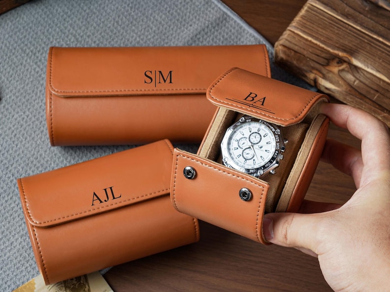 May include: Three tan leather watch rolls with snap closures and personalized monograms. One roll is open, revealing a silver-toned chronograph wristwatch with a white face inside. The monograms S|M, AJL, and BA are embossed on the exterior flaps.
