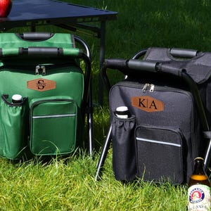 May include: A green and a black portable folding stool with an integrated cooler bag. Each bag features a brown leather-like patch with personalized lettering, a side bottle pocket, a front zippered compartment, and a sturdy black metal frame.