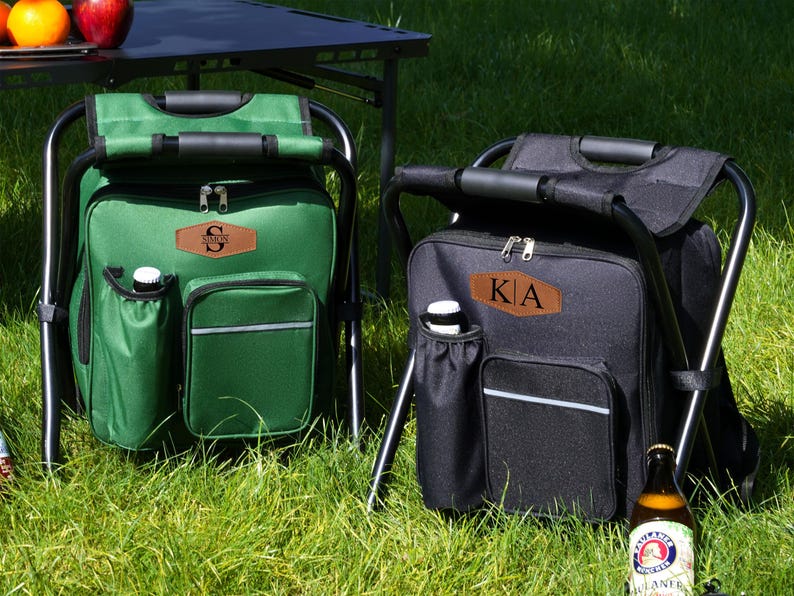 May include: A green and a black portable folding stool with an integrated cooler bag. Each bag features a brown leather-like patch with personalized lettering, a side bottle pocket, a front zippered compartment, and a sturdy black metal frame.