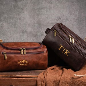May include: Two brown leather wash bags with gold zips. One bag has the name "Michael" and a mountain graphic embroidered in gold. The other bag has the initials "TIK" embroidered in gold. Both bags are on a wooden surface.