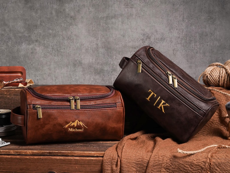 May include: Two brown leather wash bags with gold zips. One bag has the name "Michael" and a mountain graphic embroidered in gold. The other bag has the initials "TIK" embroidered in gold. Both bags are on a wooden surface.