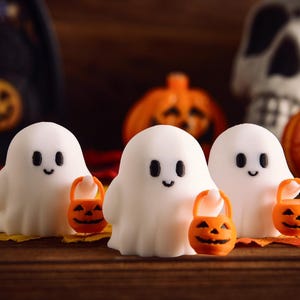 May include: Three white ghost figurines with black eyes and smiles, each holding an orange jack-o'-lantern bucket. The ghosts are on a yellow leaf, with pumpkins and a skull in the background. Halloween-themed.