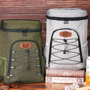 May include: Two insulated cooler backpacks, one olive green and one light gray, with black accents and front bungee cords. The olive green backpack has a brown leather patch with a "W" emblem. The gray backpack has a brown leather patch with the name "NICKY".