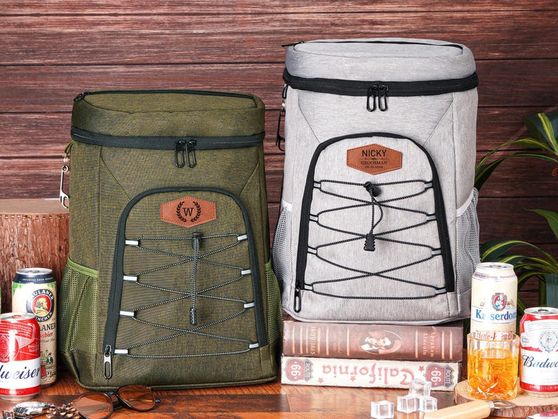 May include: Two insulated cooler backpacks, one olive green and one light gray, with black accents and front bungee cords. The olive green backpack has a brown leather patch with a "W" emblem. The gray backpack has a brown leather patch with the name "NICKY".