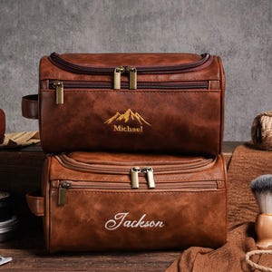 May include: Two brown leather wash bags, one on top of the other. The top bag has "Michael" embroidered below a mountain design. The bottom bag has "Jackson" embroidered on it. Both bags have gold zips.