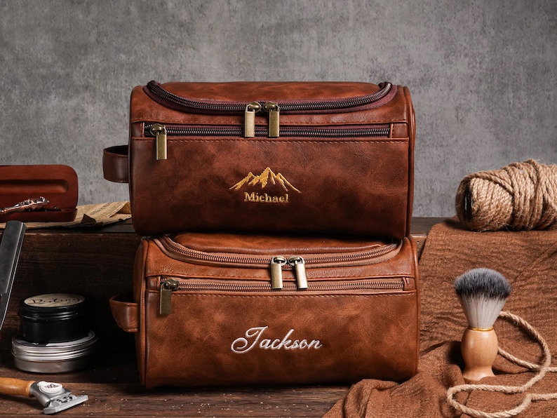 May include: Two brown leather wash bags, one on top of the other. The top bag has "Michael" embroidered below a mountain design. The bottom bag has "Jackson" embroidered on it. Both bags have gold zips.