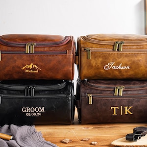 May include: Four personalised leather wash bags in shades of brown and black. Each bag has a zip closure and a handle. The bags are embossed with names, initials, and a mountain graphic. The text "GROOM 08.06.25" is also visible.